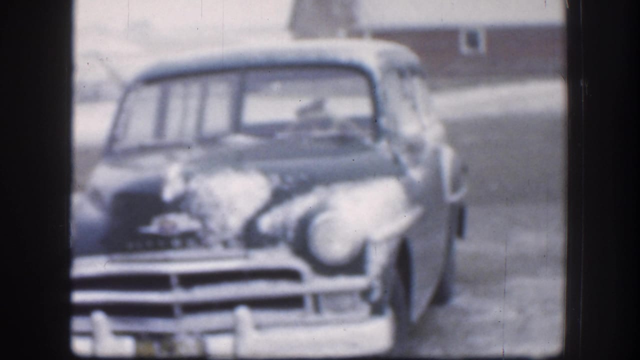 Close front-on 8mm shot of a snow-laden 1950s American sedan on a Kansas farm, circa 1958; outbuilding faintly visible behind.