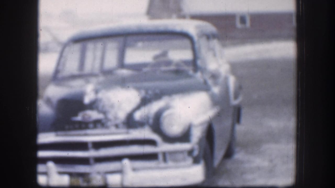 Near-identical front view of snow-laden 1950s sedan; chrome grille prominent, farm structure faintly visible in background.