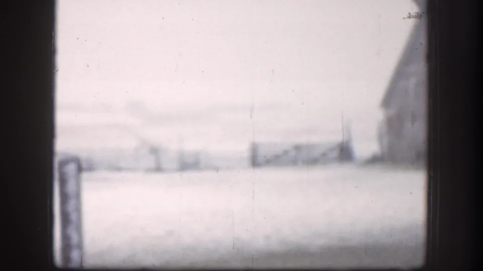 Wide 8mm exterior of a Kansas farm in blizzard conditions, circa 1958; barn mid-distance across a snow-covered field with fence posts in foreground.