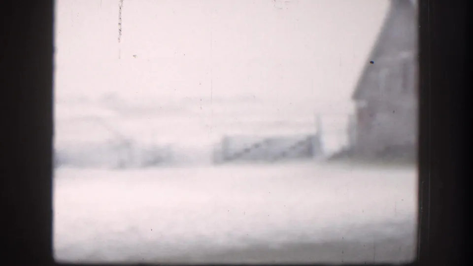 Wide 8mm exterior of a Kansas farm during a blizzard, circa 1958; barn at right edge, fence line across foreground, near-whiteout conditions throughout.