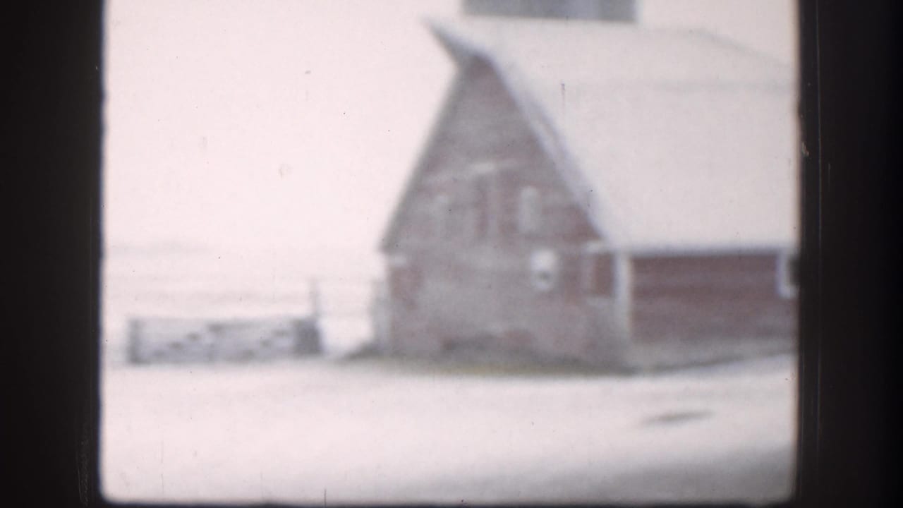 8mm home movie frame, circa 1958, showing a snow-covered Kansas farmyard with a red barn and wooden outbuildings under an overcast winter sky.