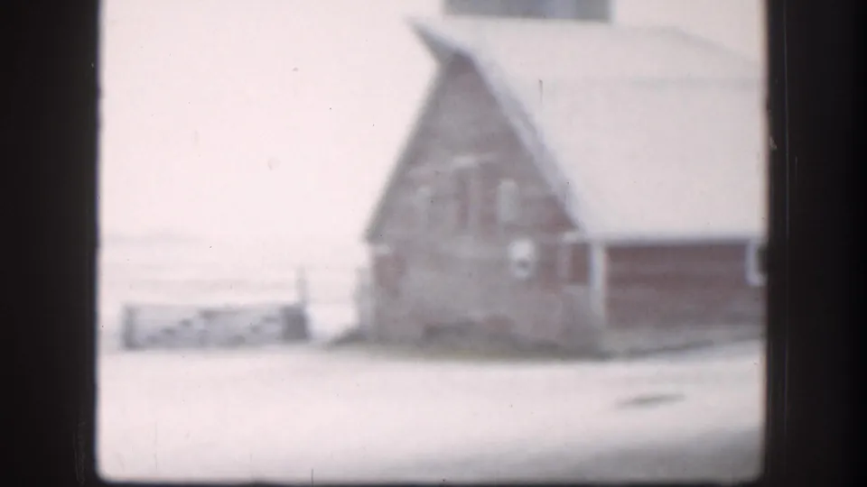 8mm home movie frame, circa 1958, showing a snow-covered Kansas farmyard with a red barn and wooden outbuildings under an overcast winter sky.