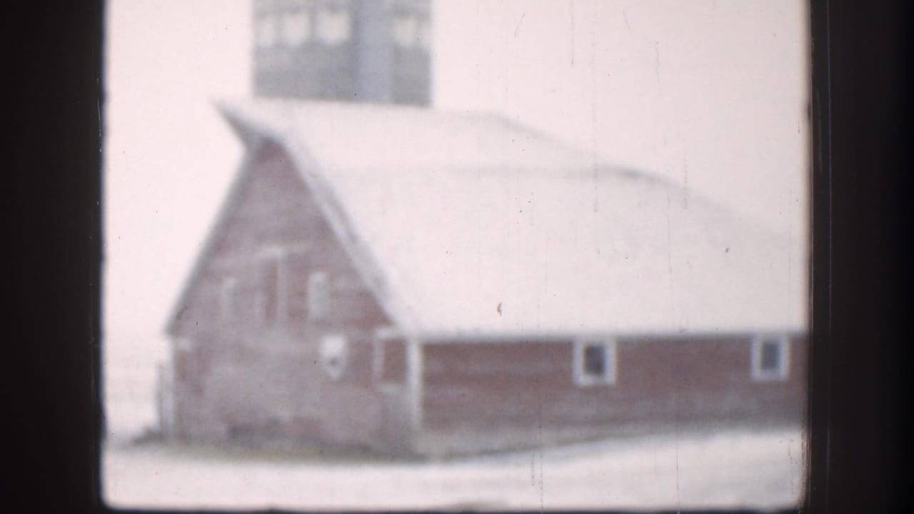 8mm home movie frame, circa 1958, of a red barn with steeply pitched snow-covered roof and adjacent outbuildings on a Kansas farm in winter.