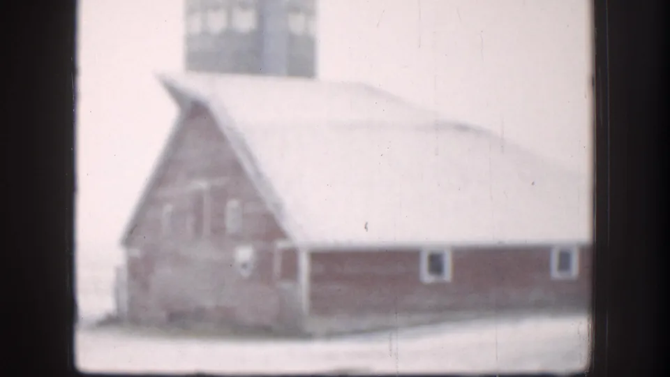 8mm home movie frame, circa 1958, of a red barn with steeply pitched snow-covered roof and adjacent outbuildings on a Kansas farm in winter.