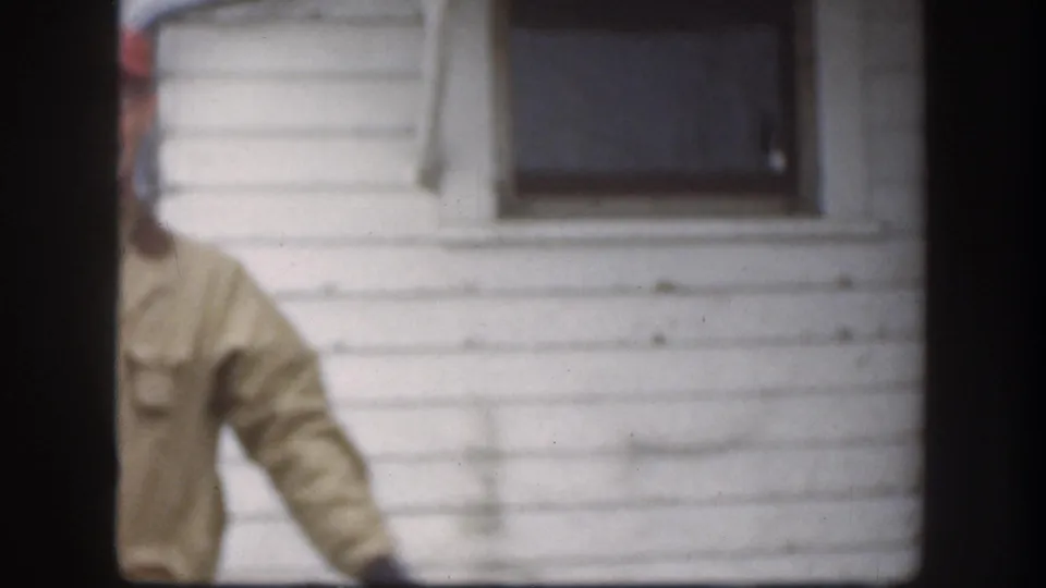 8mm home movie frame, circa 1958, of an adult man in a tan work jacket before a white clapboard farm building; soft focus is characteristic of 8mm film.