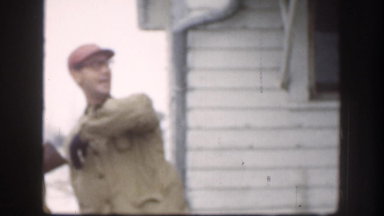 Man in red baseball cap and beige jacket gesturing outdoors beside a white wood-sided house.