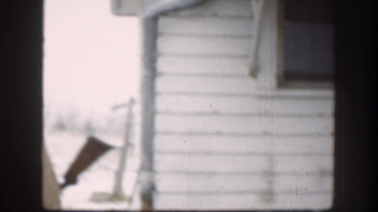 White clapboard house exterior with dark elongated prop object visible, 1958 8mm home movie, Kansas USA.