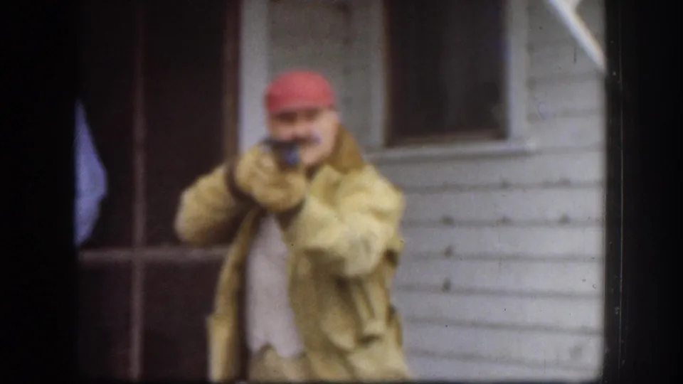 Man in red cap and tan jacket raising a small dark prop toward the camera outside a white clapboard house, circa 1958 Kansas USA, 8mm home movie.