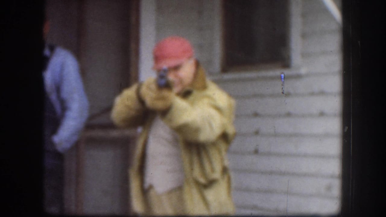 Man in a red hunting cap and olive field coat aims a hunting rifle at the camera in a candid 8mm home movie moment, consistent with Kansas circa 1958.