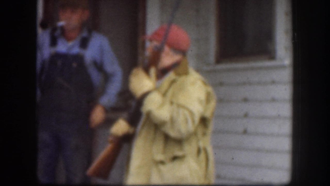 Two adult men outside a clapboard house in what appears to be a late-1950s winter setting — one in a red hunting cap and olive coat holds a rifle at his side, the other in denim bib overalls.