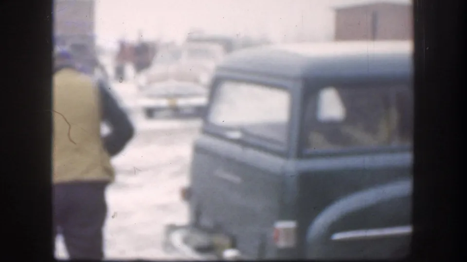 Man in olive field coat moves toward dark teal rounded-body American cars in a snowy residential yard, with background figures visible, consistent with a late-1950s Midwestern winter scene.