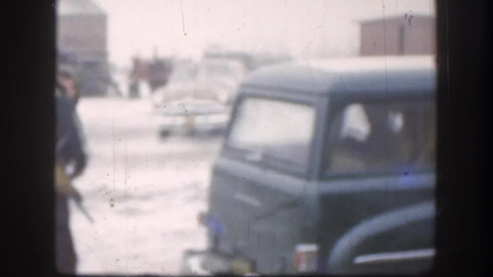 Dark teal rounded-body American cars dominate the foreground of a snow-covered residential yard; background figures and strong snow glare are consistent with late-1950s 8mm home movie footage.