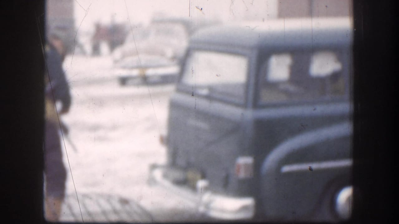 A dark-blue late-1950s station wagon sits in a snow-covered open lot in Kansas, 1958; a person in dark winter clothing stands partially cropped at the left edge.