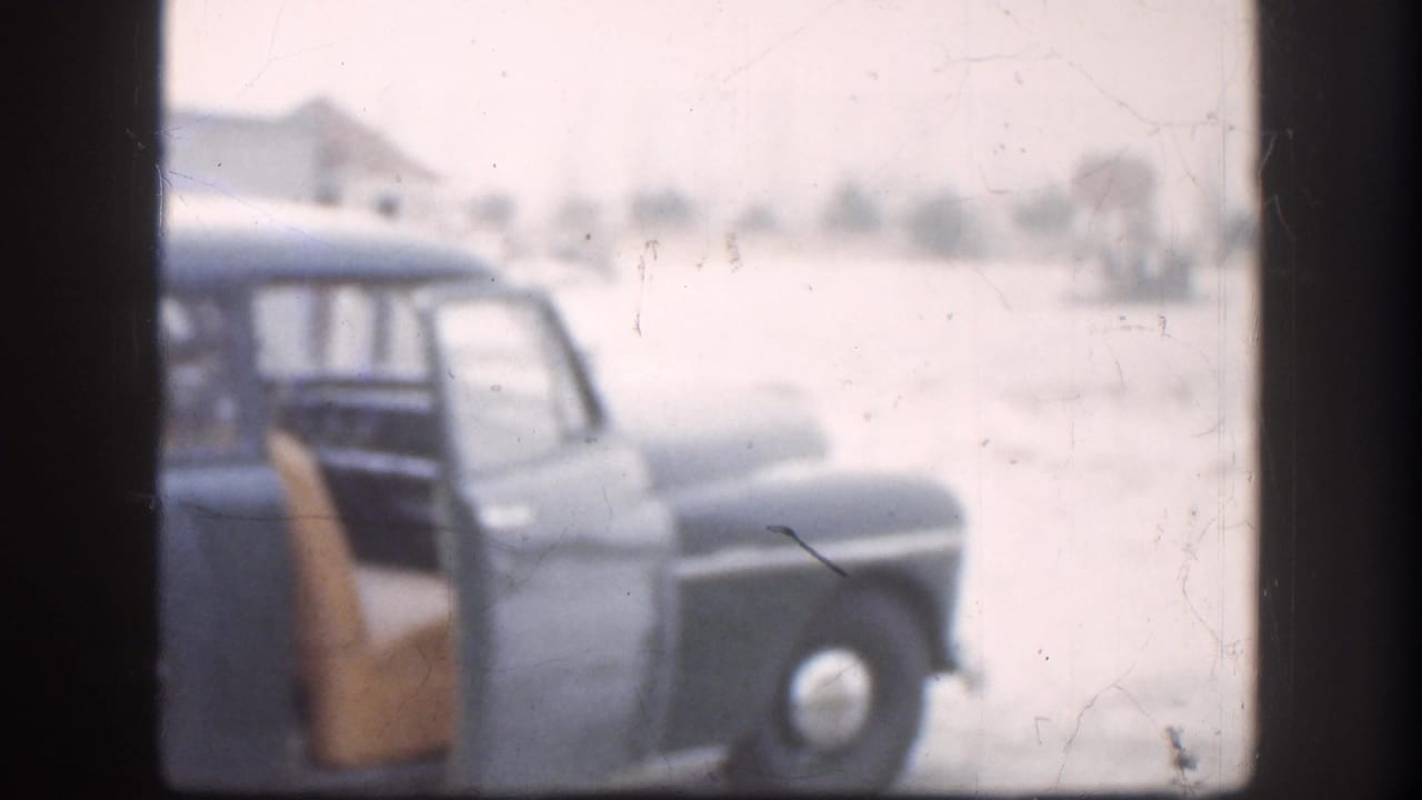 A late-1950s dark-blue station wagon with its rear door open stands in a snow-covered lot in what appears to be Kansas, 1958; the flat Great Plains horizon stretches behind it.