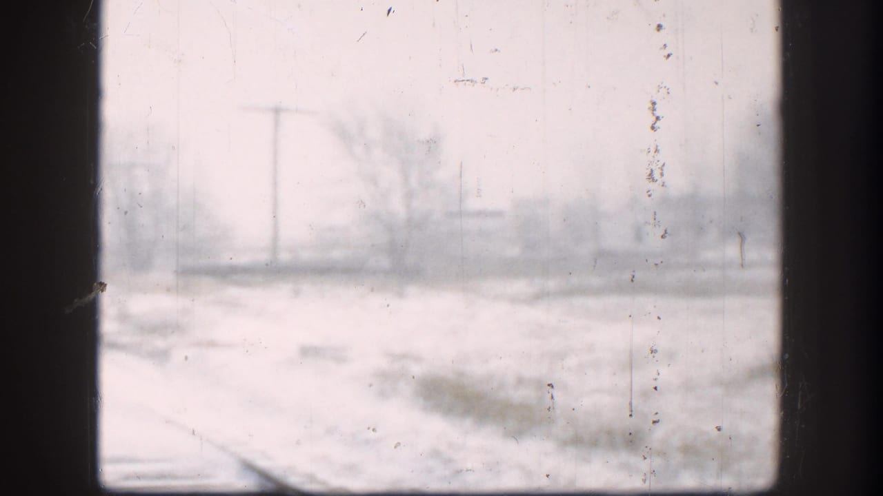 8mm frame showing Kansas railroad tracks disappearing into heavy snowfall, captured through a grime-coated window, circa 1958.