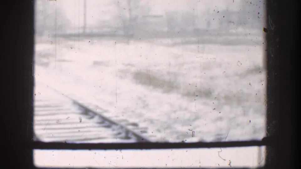 Close view of snow-dusted railroad tracks through a window, Kansas circa 1958, with flat snow-covered terrain extending into blizzard conditions.