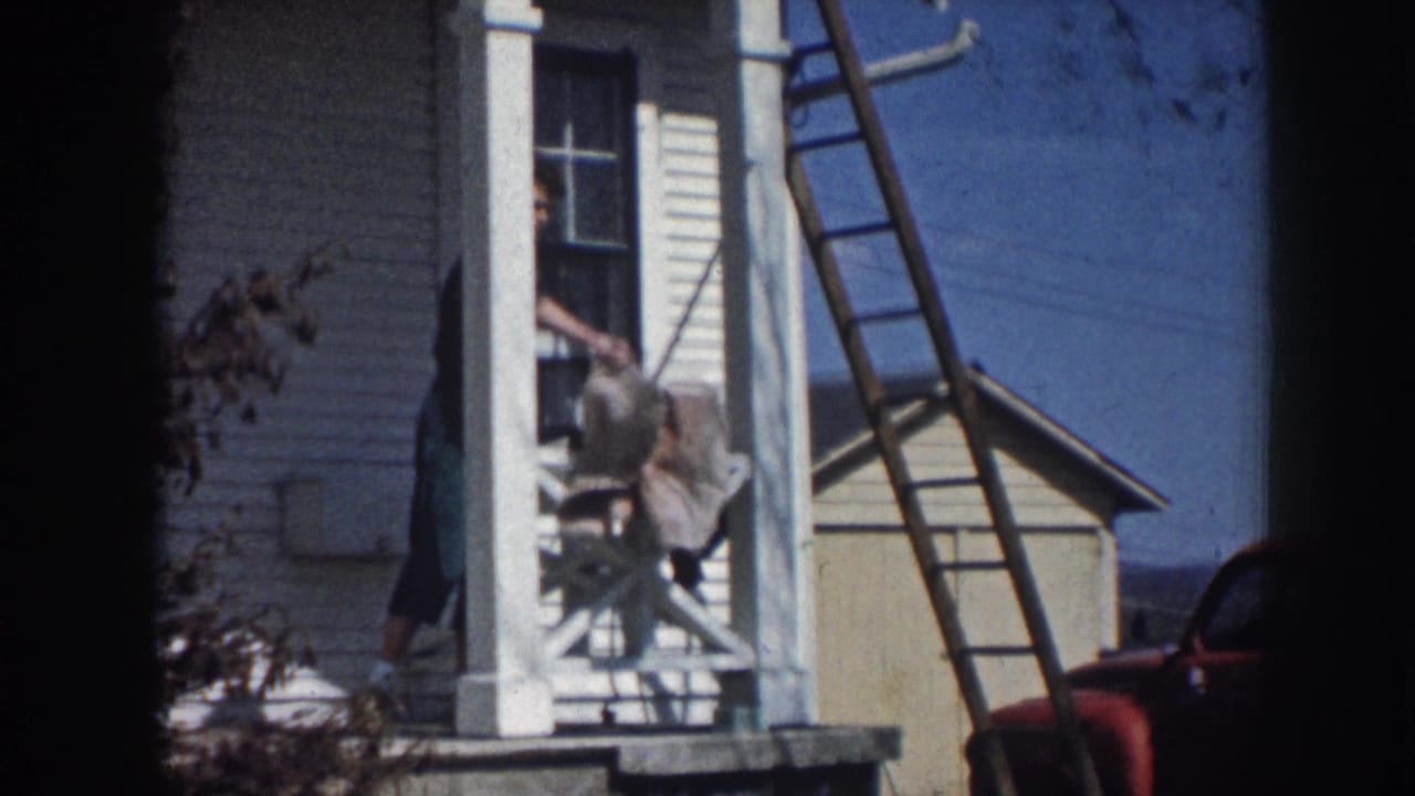 Figure descending porch steps of white clapboard farmhouse with ladder against the wall, consistent with rural Nebraska circa 1958; red vehicle partially visible.