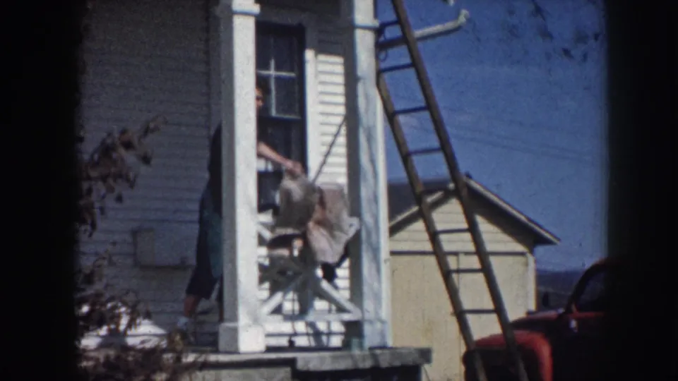 Figure descending porch steps of white clapboard farmhouse with ladder against the wall, consistent with rural Nebraska circa 1958; red vehicle partially visible.