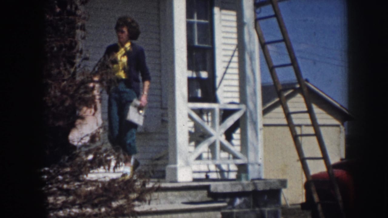 Woman in yellow shirt and dark cardigan carrying papers or a bundle down porch steps of white clapboard farmhouse, consistent with rural Nebraska circa 1958; ladder against house.
