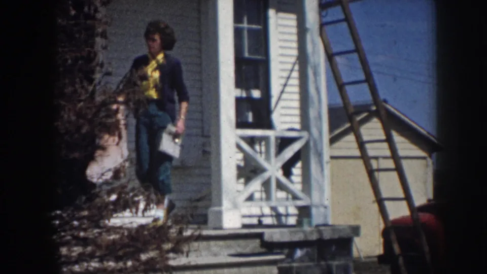 Woman in yellow shirt and dark cardigan carrying papers or a bundle down porch steps of white clapboard farmhouse, consistent with rural Nebraska circa 1958; ladder against house.