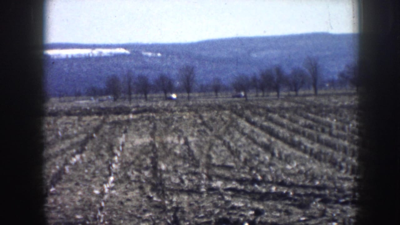 8mm home movie still of plowed agricultural fields, bare deciduous tree line, and low hills under a pale sky, consistent with rural Nebraska circa 1958.