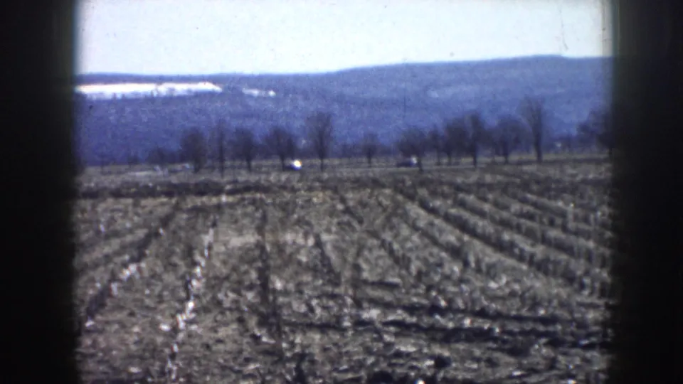 8mm home movie still of plowed agricultural fields, bare deciduous tree line, and low hills under a pale sky, consistent with rural Nebraska circa 1958.