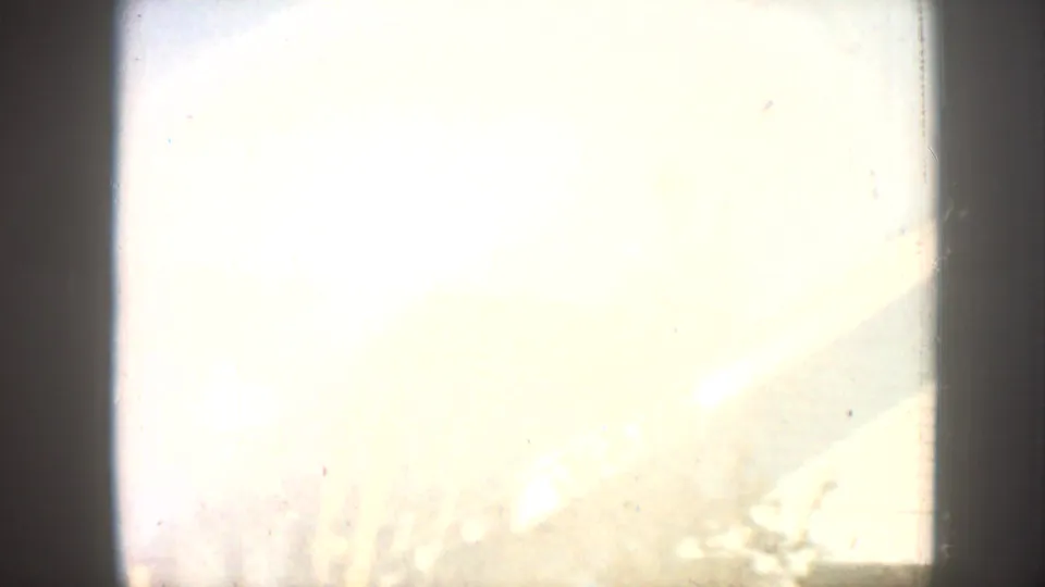 Overexposed 8mm film frame showing a wash of blown-out light with a barely visible structural edge at lower right; likely a scene transition.