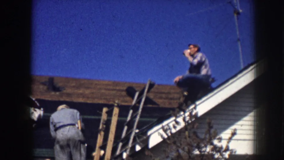 8mm home movie still of two adult men on a residential roofline — one seated at the peak drinking from a cup, the other at the base of an extension ladder, Nebraska circa 1958.