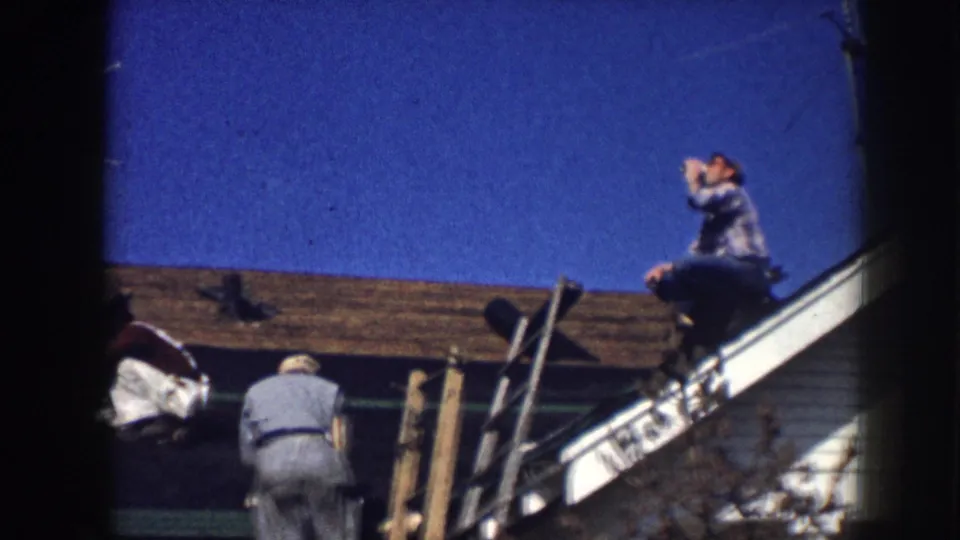 8mm home movie continuation showing the same two men on a residential roofline — one seated at the peak with cup raised, the other at the ladder — under a vivid blue sky, Nebraska circa 1958.