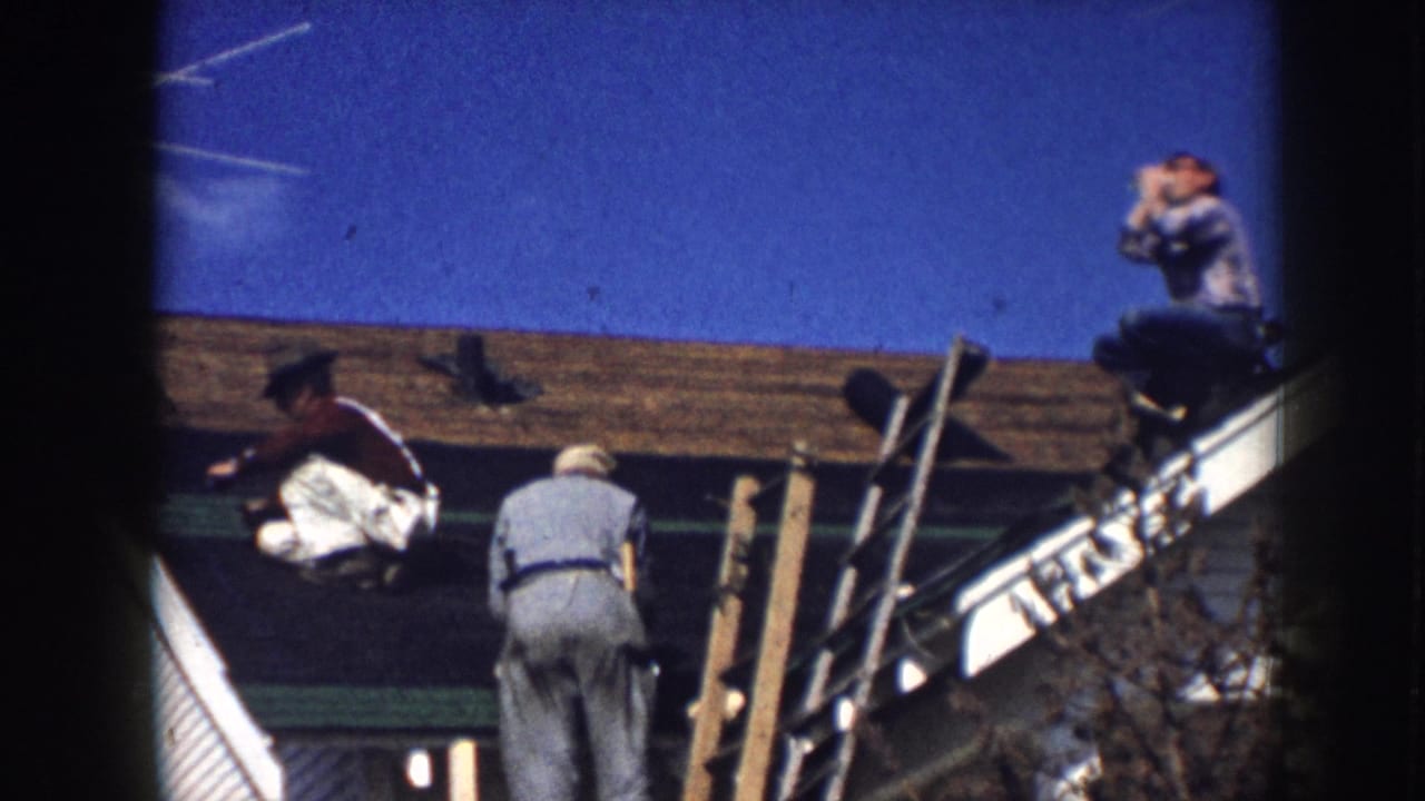 Three men re-roofing a clapboard house in what appears to be Nebraska, USA, circa 1958; one rests at the ridge while two others work near wooden pole scaffolding.