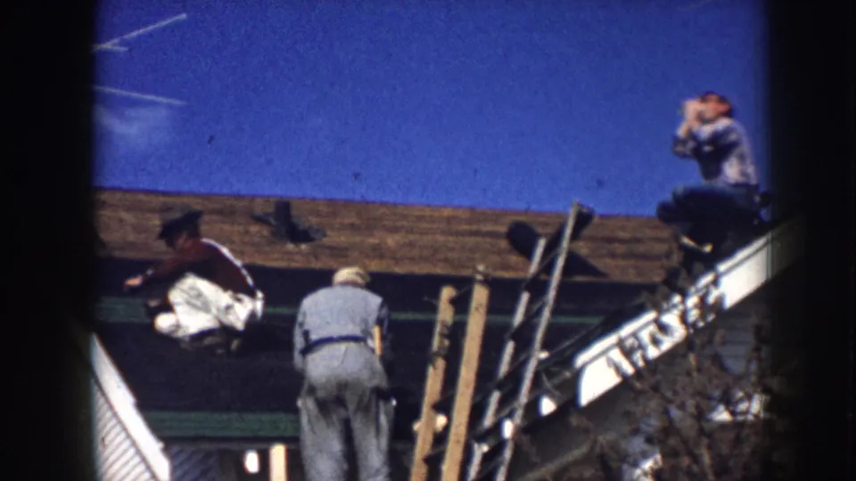 Three men re-roofing a clapboard house in what appears to be Nebraska, USA, circa 1958; one rests at the ridge while two others work near wooden pole scaffolding.