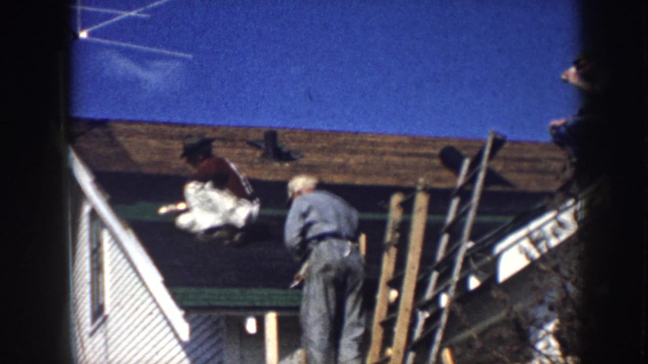 Two roofers working on a clapboard house exterior circa 1958, with wooden pole scaffolding, exposed roof sheathing, and a deep blue sky visible.