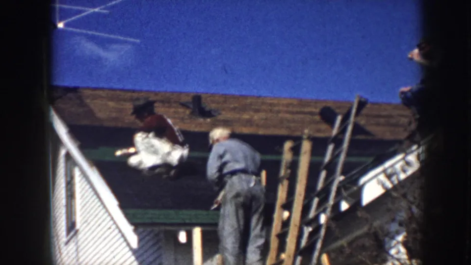 Two roofers working on a clapboard house exterior circa 1958, with wooden pole scaffolding, exposed roof sheathing, and a deep blue sky visible.