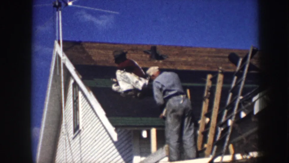 Close view of a worker in 1950s work clothes handling bundled roofing material at the eave of a clapboard house, wooden pole scaffolding in the background.