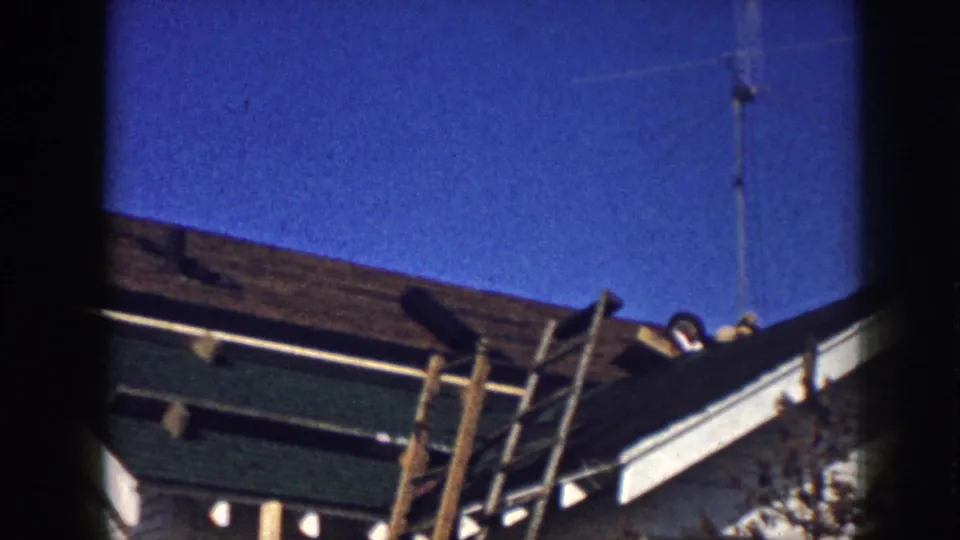 Wide shot of a mid-century residential re-roofing job with exposed sheathing, wooden scaffolding, and workers partly visible on the slope against a blue sky, circa 1958.