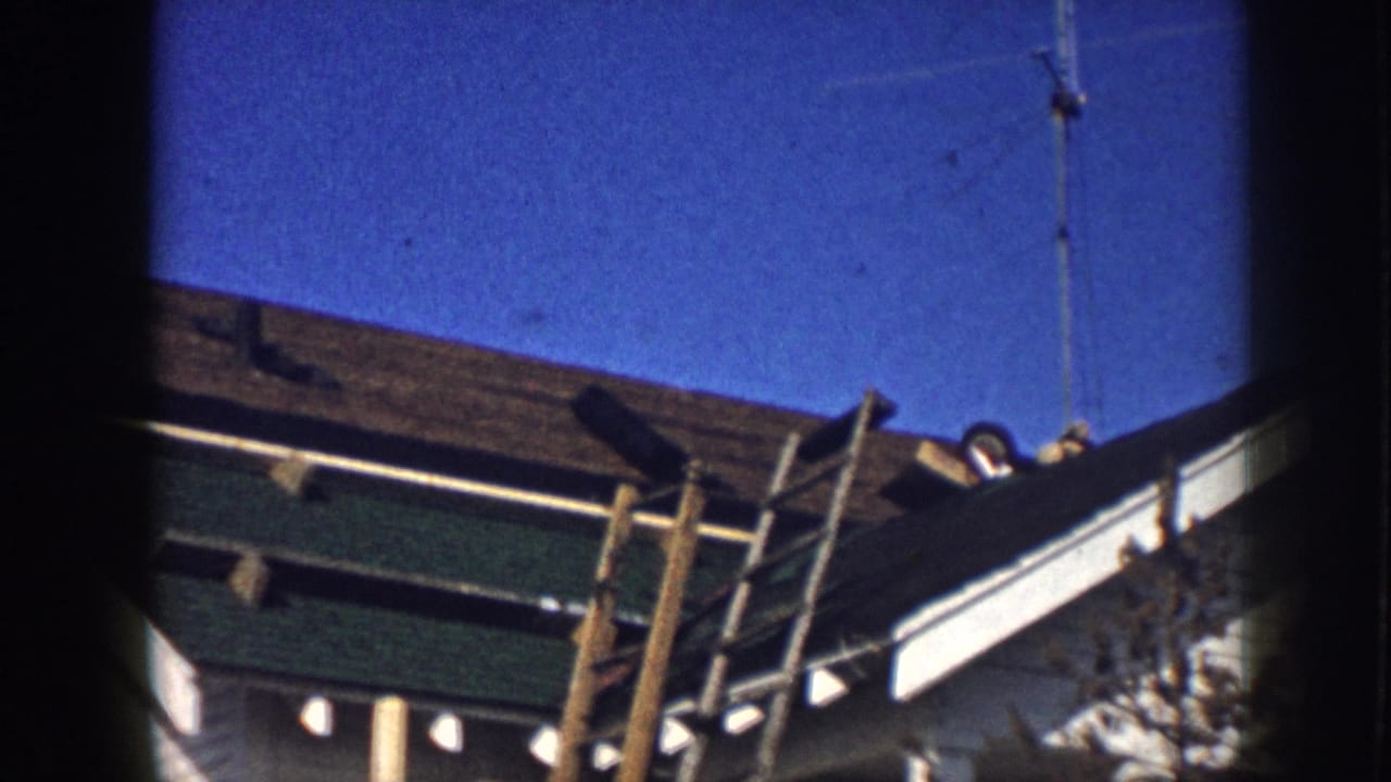 Low-angle 8mm shot of a residential roofline in Nebraska, circa 1958, showing a wooden ladder, green fascia trim, and an antenna pole against a clear blue sky.