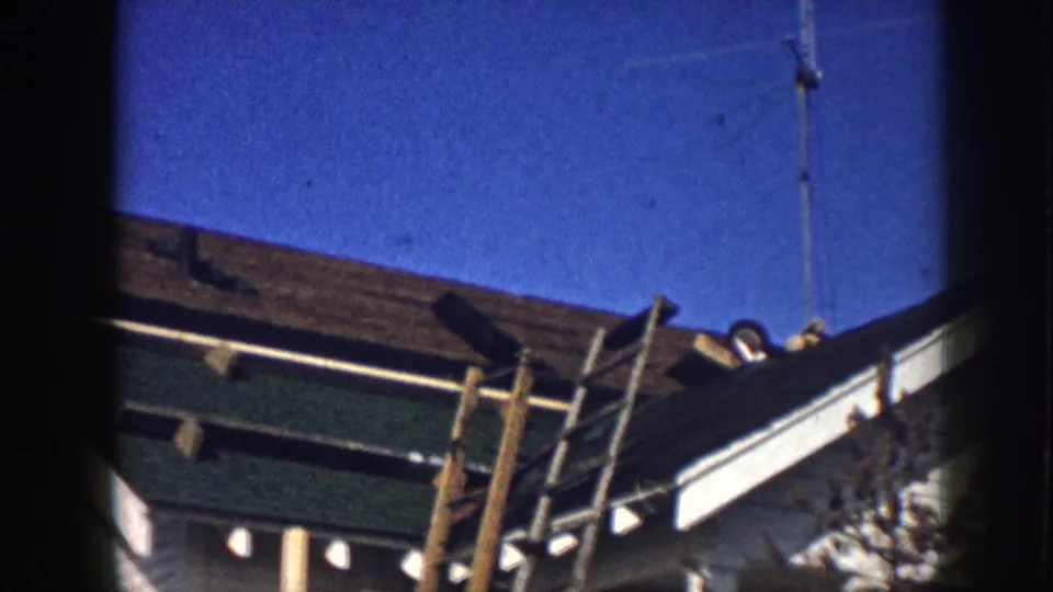 Low-angle 8mm shot of a residential roofline in Nebraska, circa 1958, showing a wooden ladder, green fascia trim, and an antenna pole against a clear blue sky.