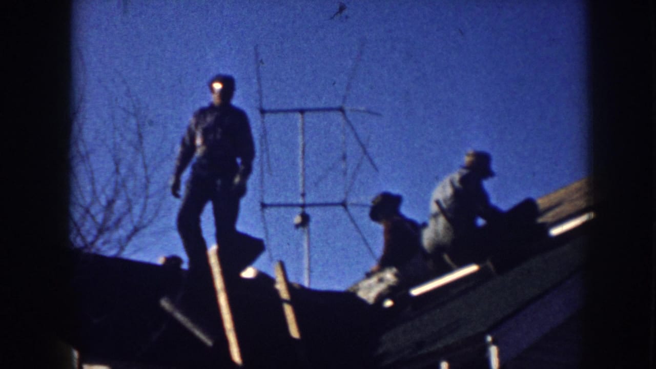 Two men silhouetted on a rooftop alongside a yagi television antenna against a vivid blue sky, with bare tree branches at upper left.