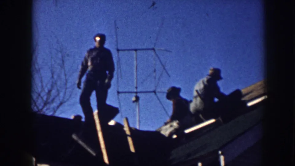 Two men silhouetted on a residential rooftop in Nebraska, circa 1958, positioning a yagi TV antenna under a clear blue 8mm sky, with bare tree branches in the background.