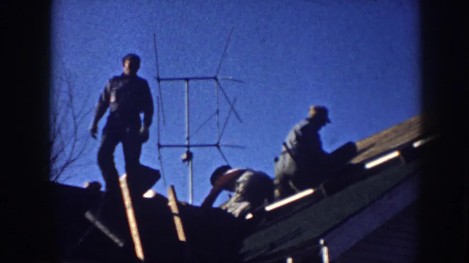 Two men silhouetted on a residential rooftop in Nebraska, circa 1958, one standing and one crouching while working on a yagi TV antenna, against a deep blue 8mm sky.