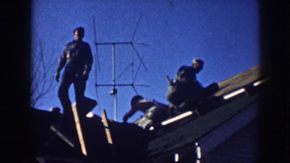 Two men in period work clothing silhouetted on a residential rooftop in Nebraska, circa 1958, alongside a yagi TV antenna under a clear blue 8mm sky.