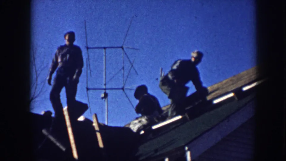 Three men working on a sloped shingle roof in Nebraska, c.1958, silhouetted against a vivid blue sky with a rooftop Yagi TV antenna behind them.