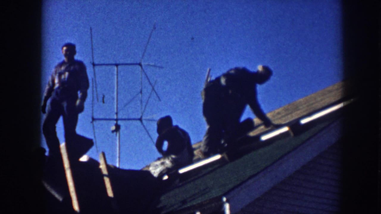 Three men on a roof in Nebraska, c.1958; one standing, two actively working, silhouetted against a saturated blue sky with a Yagi TV antenna behind them.