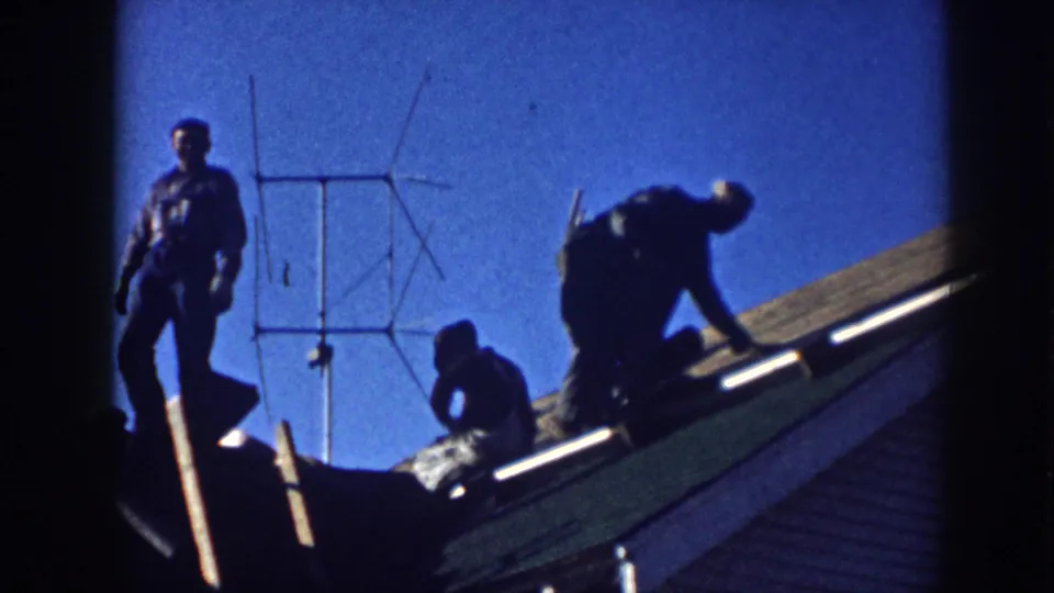 Three men on a roof in Nebraska, c.1958; one standing, two actively working, silhouetted against a saturated blue sky with a Yagi TV antenna behind them.