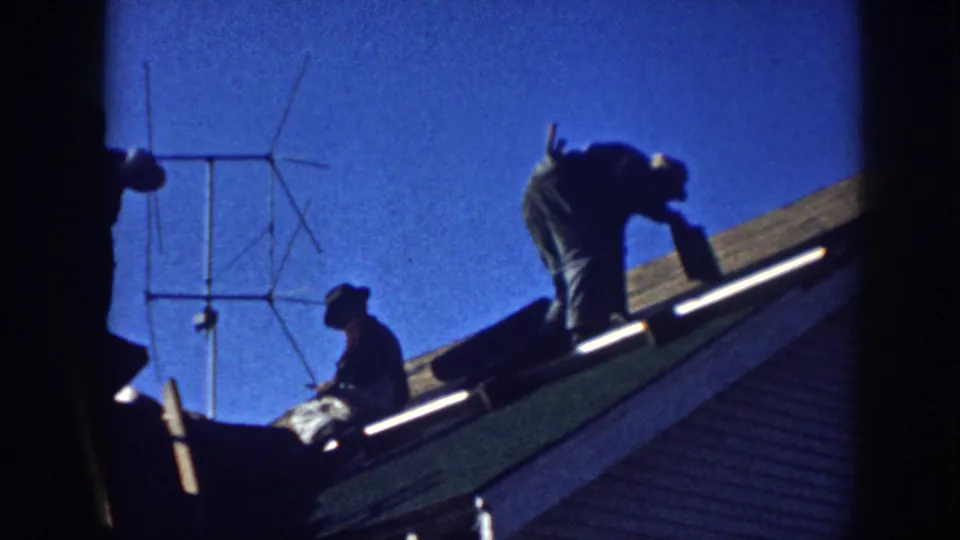 Two men on a sloped shingled roof in Nebraska, c.1958; one bent at work with a Yagi-type TV antenna to the left against a vivid blue sky.