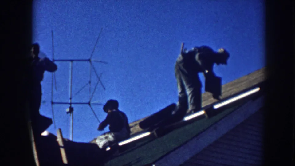 Two men on a sloped shingled roof in Nebraska, c.1958; one crouching at work with a Yagi-type TV antenna to the left and a vivid blue sky above.