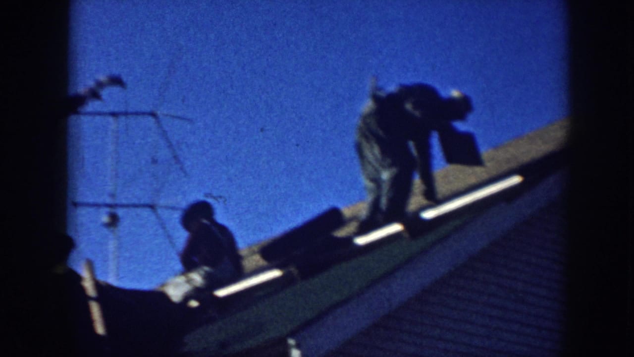 Two men silhouetted on a residential roof slope against a deep blue sky in what appears to be late-1950s Nebraska; TV antenna and roofing materials visible.
