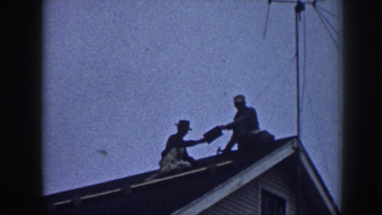 Two silhouetted figures near a roof ridge, one appearing to pass material to the other, TV antenna upper right, clapboard siding and window below.