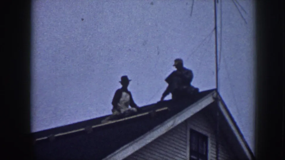 Two men on a suburban gabled roof circa 1958, one in a brimmed hat; the gable peak, clapboard siding, and a window frame the scene against a pale blue-grey sky.