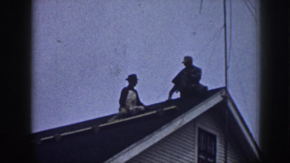 Two men on a suburban gabled roof circa 1958, one in a brimmed hat; composition nearly identical to preceding frame, suggesting a static handheld camera with minimal movement.
